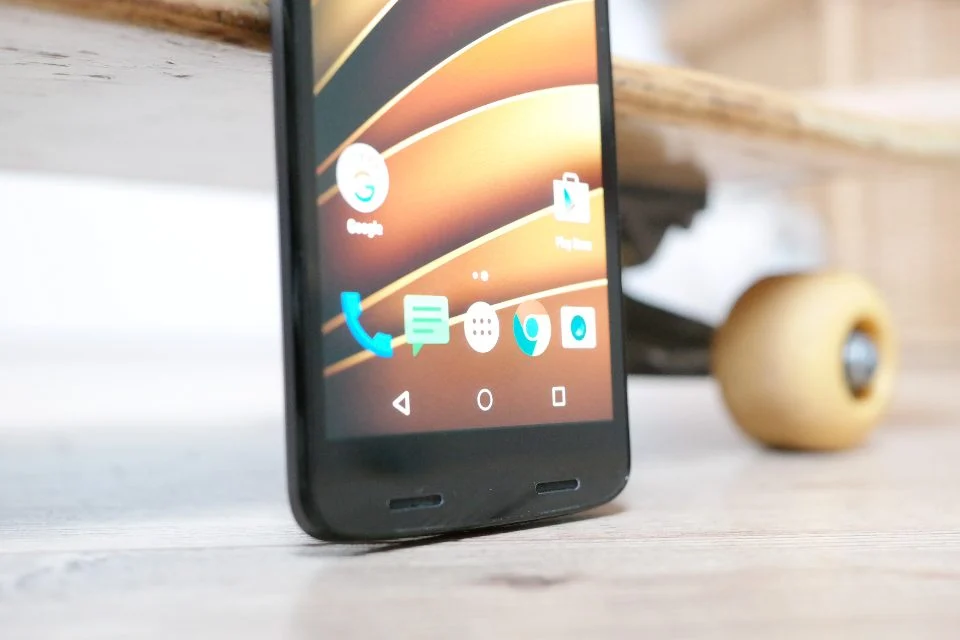 Celular Android com a tela inicial, apoiado no chão ao lado da roda de um skate