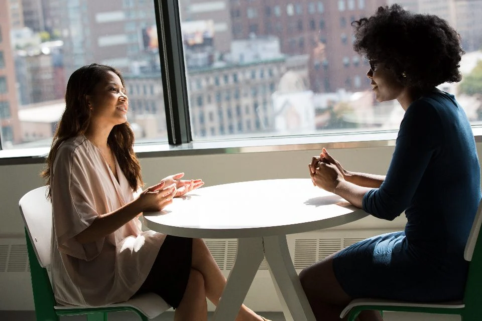 Duas mulheres conversando sentadas a uma mesa, perto de uma janela