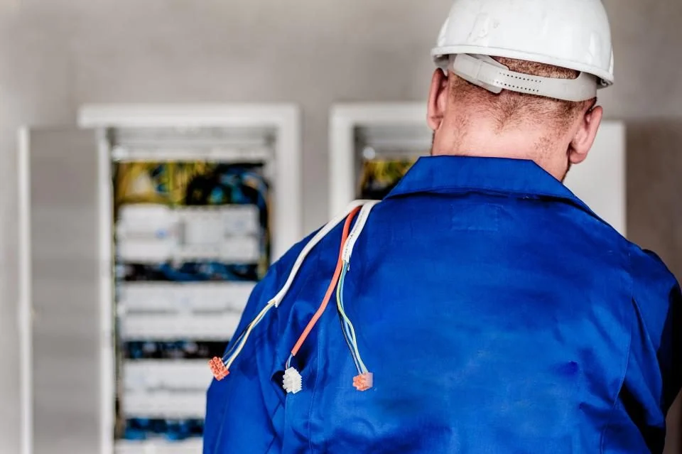 Eletricista de capacete diante de um quadro de distribuição elétrica aberto