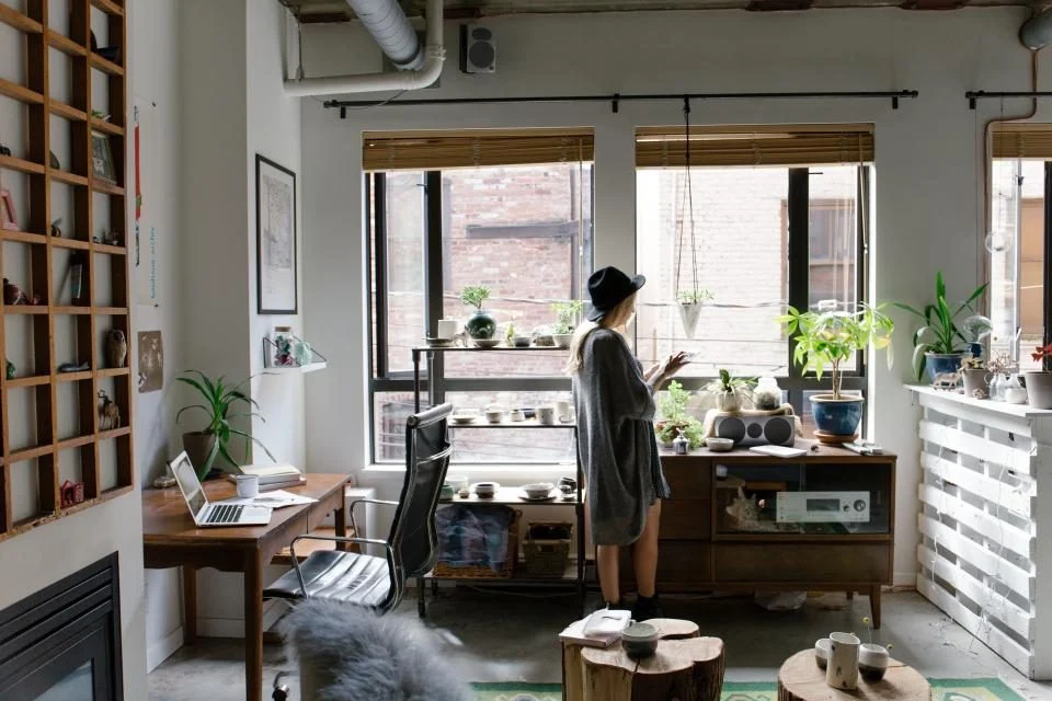 Escritório em casa com mesa de trabalho, estantes, plantas e uma pessoa perto da janela