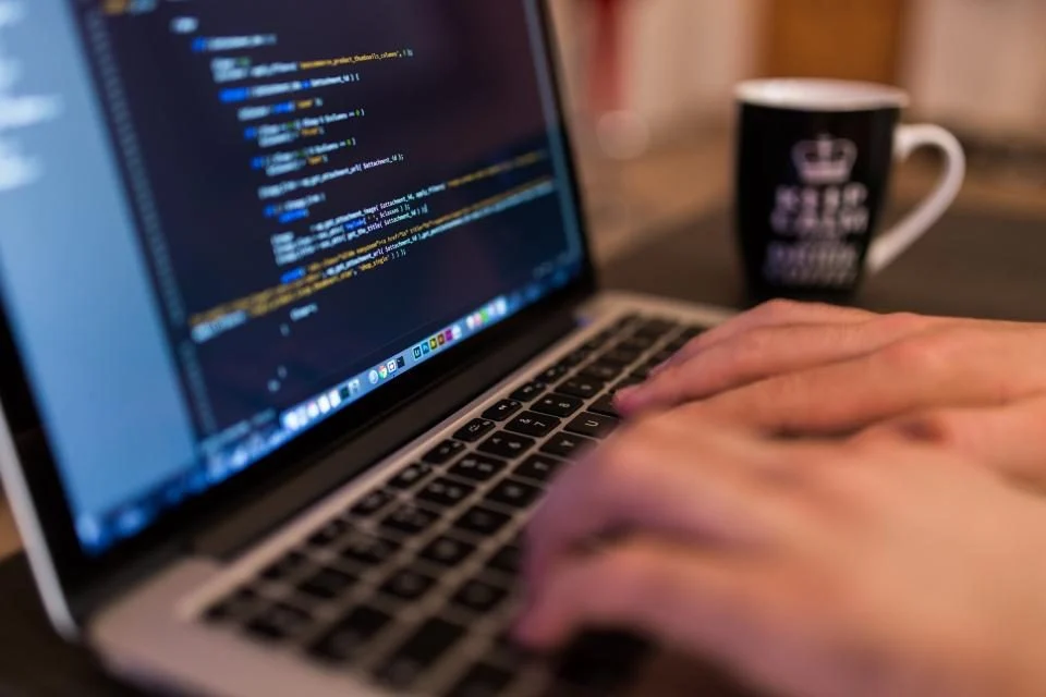 Mãos digitando num notebook com código na tela, ao lado de uma caneca