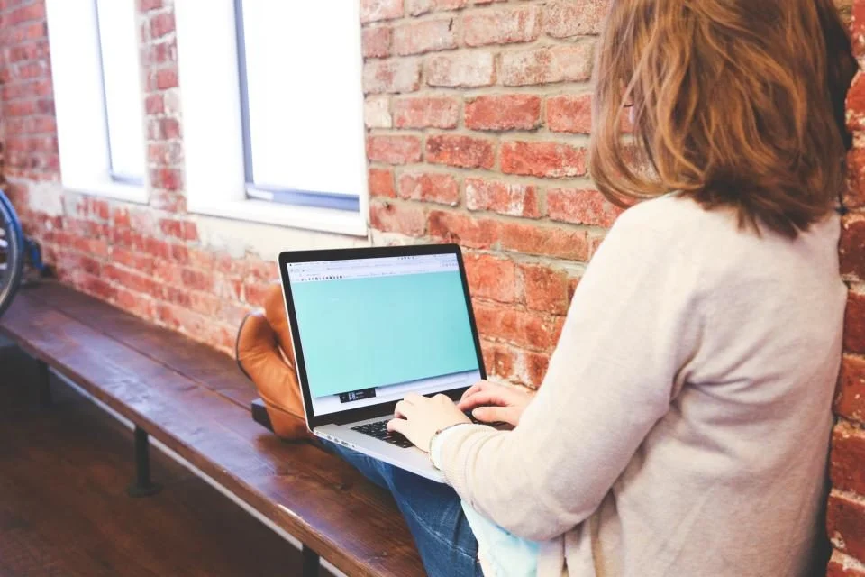 Mulher digitando num notebook sentada num banco, junto a uma parede de tijolos